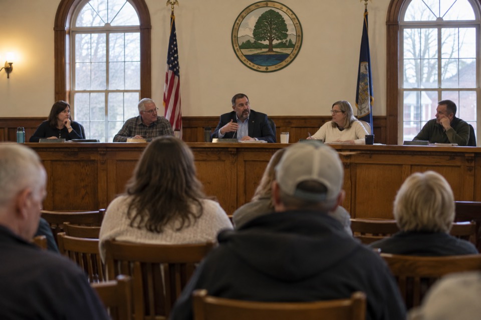 Five trustees seated at the dais during a public meeting while residents watch from the audience